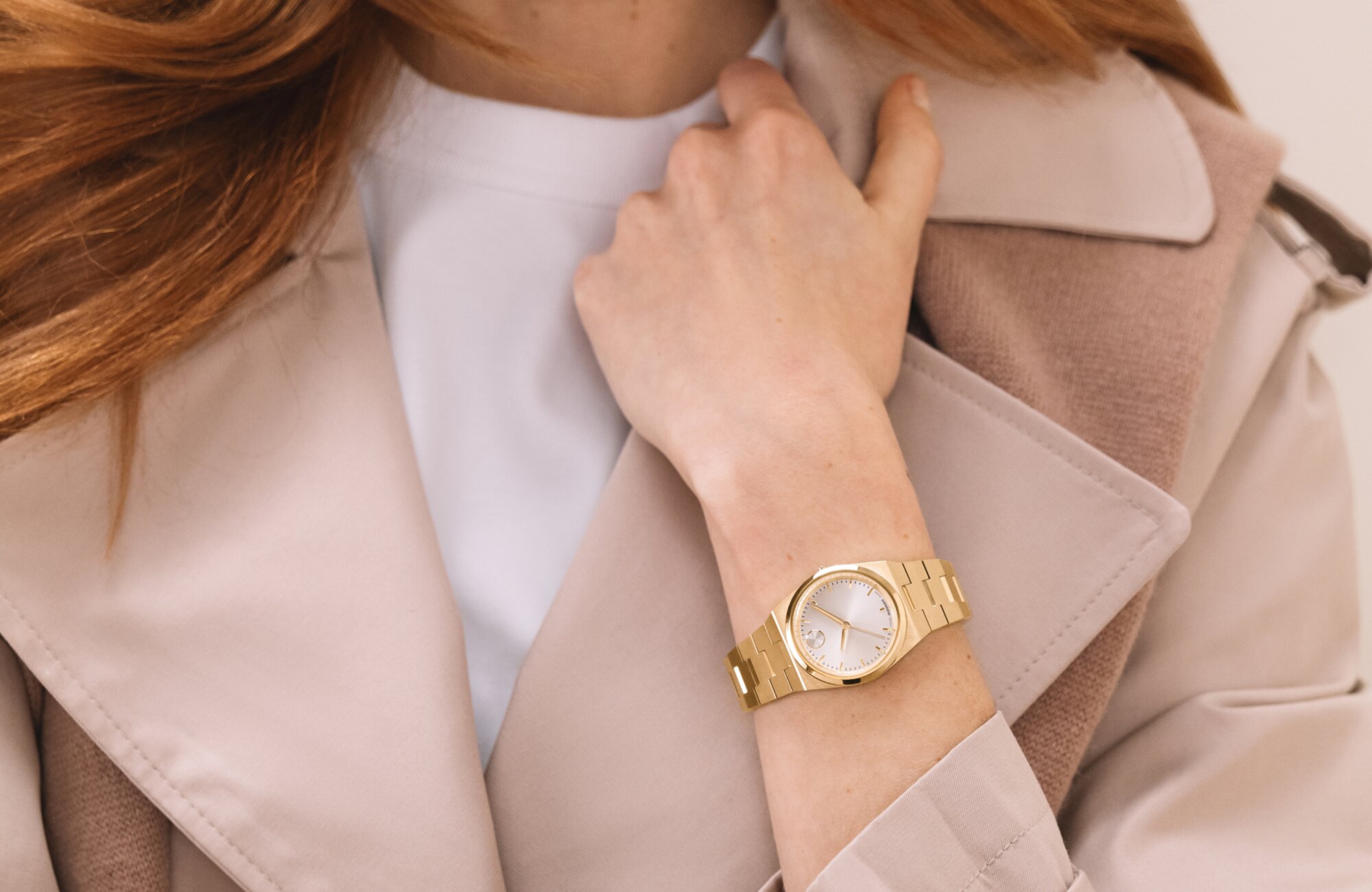 Woman in a pink coat and white t-shirt, holding her collar, wearing the gold tone BOLD Quest Mini bracelet watch.