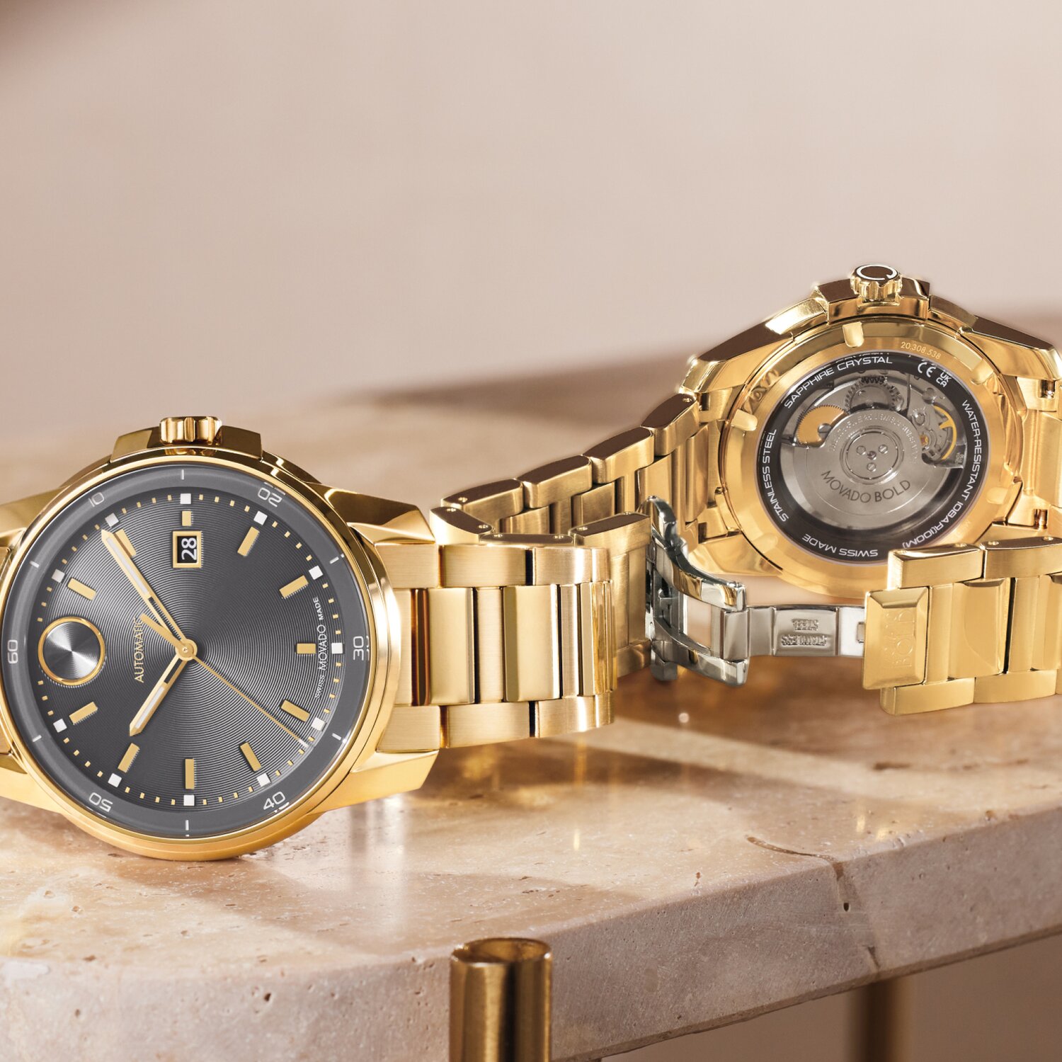 Front of a BOLD Verso Automatic watch with grey dial and gold tone bracelet laying in front of the back of the BOLD Verso Automatic watch with a caseback that displays the automatic movement