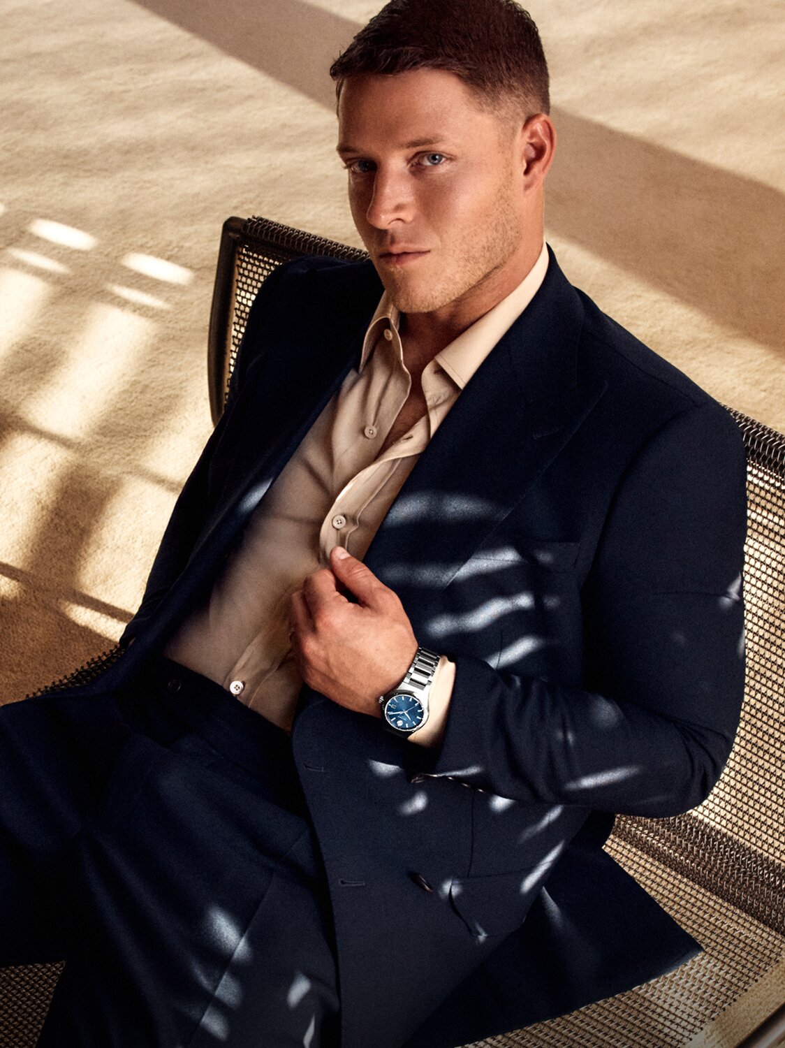 Christian McCaffrey sitting on a chair, holding his suit jacket and wearing the Museum Imperiale watch with stainless steel bracelet and blue dial.