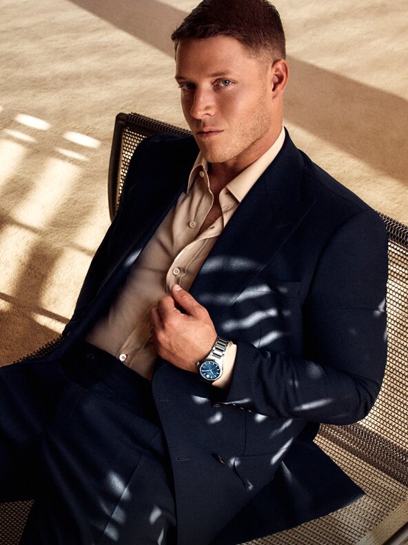 Christian McCaffrey sitting on a chair, holding his suit jacket and wearing the Museum Imperiale watch with stainless steel bracelet and blue dial.