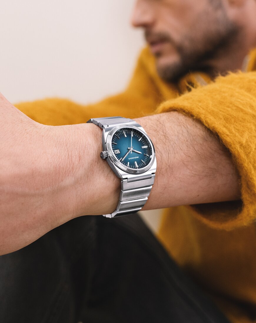 Museum Classic Automatic watch with stainless steel bracelet and black dial in front of a yellow background.