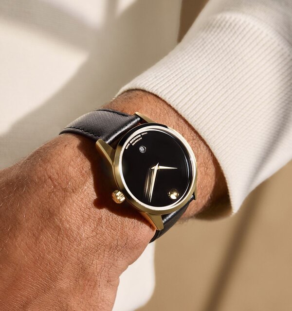 Man in a white sweater wearing the black Museum Classic automatic strap watch with gold tone accents.