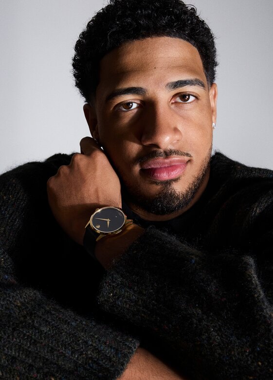 Tyrese Haliburton sitting in front of a grey background wearing a black sweater and the Museum Classic automatic watch with black strap and dial with gold accents.