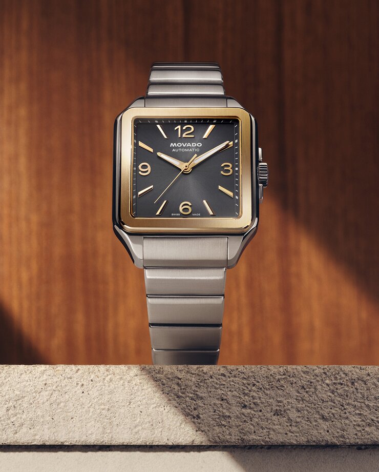 Heritage 1917 watch with stainless steel bracelet and grey dial sitting on stone in front of a wooden wall.