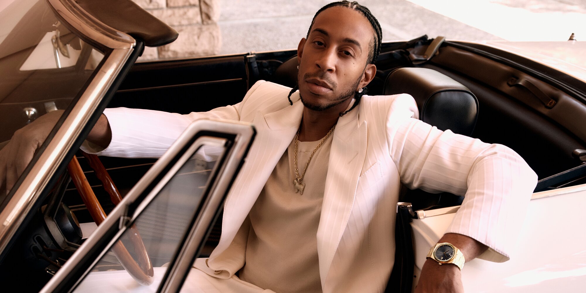 Ludacris wearing a white suit, sitting in a car, wearing the BOLD Quest watch with gold tone bracelet and black dial.