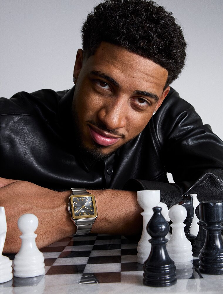 Tyrese Haliburton leaning a chess set in a black leather jacket while wearing the Heritage 1917 watch with stainless steel bracelet and grey dial.
