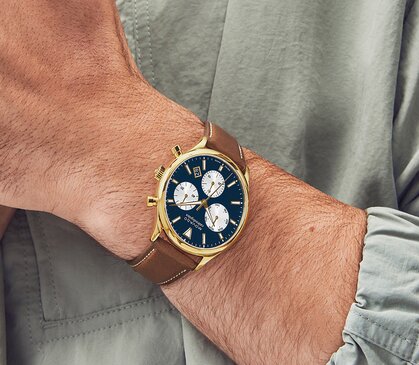 Man in a green jacket wearing the Heritage Calendoplan watch with brown strap and blue chronograph dial.
