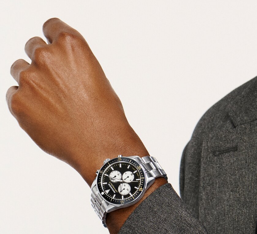Man in a grey coat wearing the Heritage Calendoplan S watch with stainless steel bracelet and black chronograph dial.