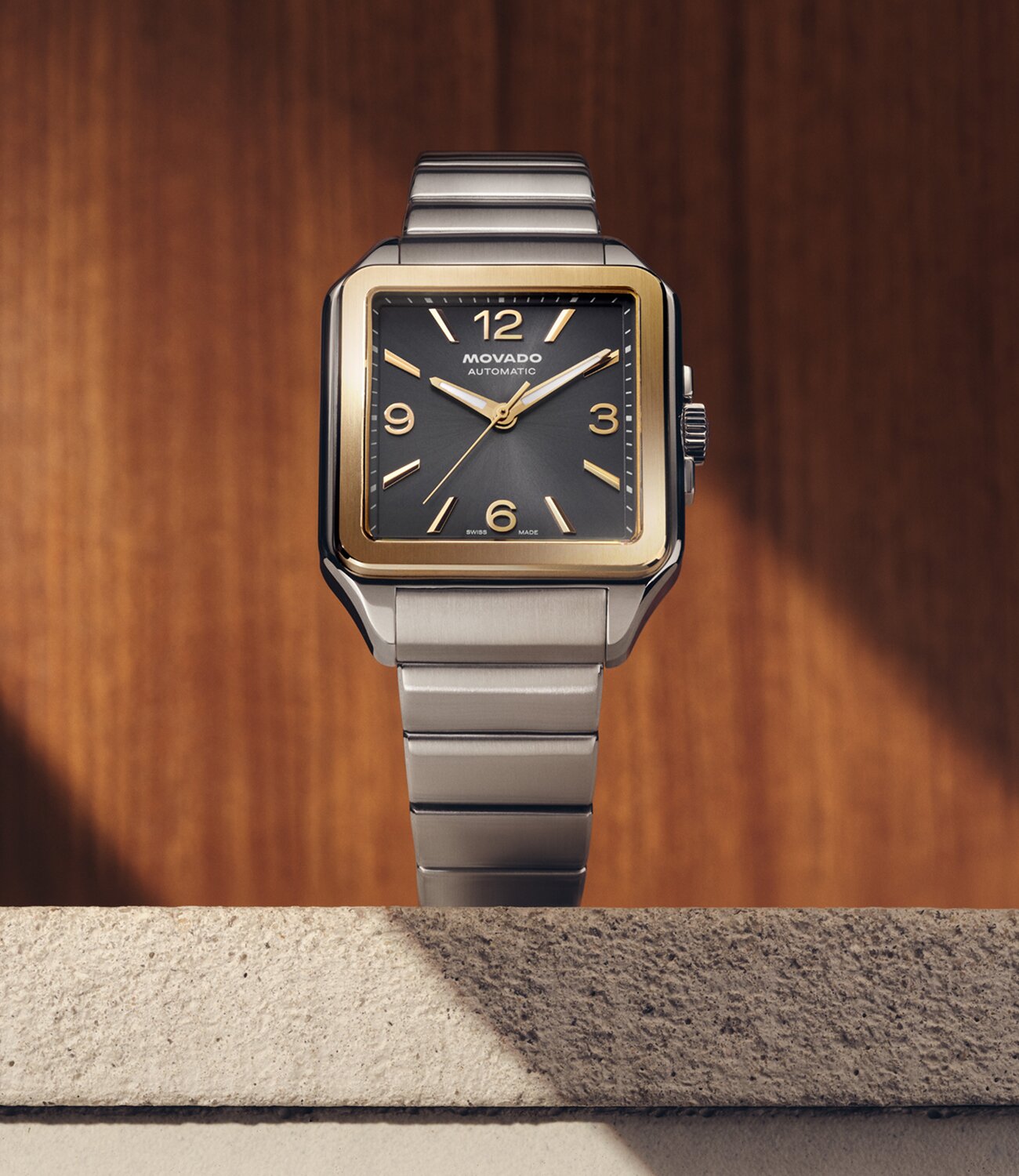 Heritage 1917 watch with stainless steel bracelet and grey dial sitting on stone in front of a wooden wall.