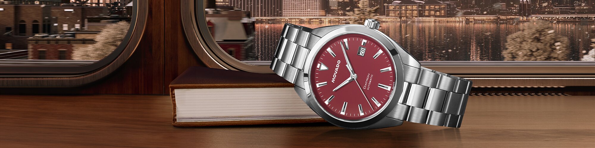 Silver watch with red dial laying on a book on a train with a cityscape in the background