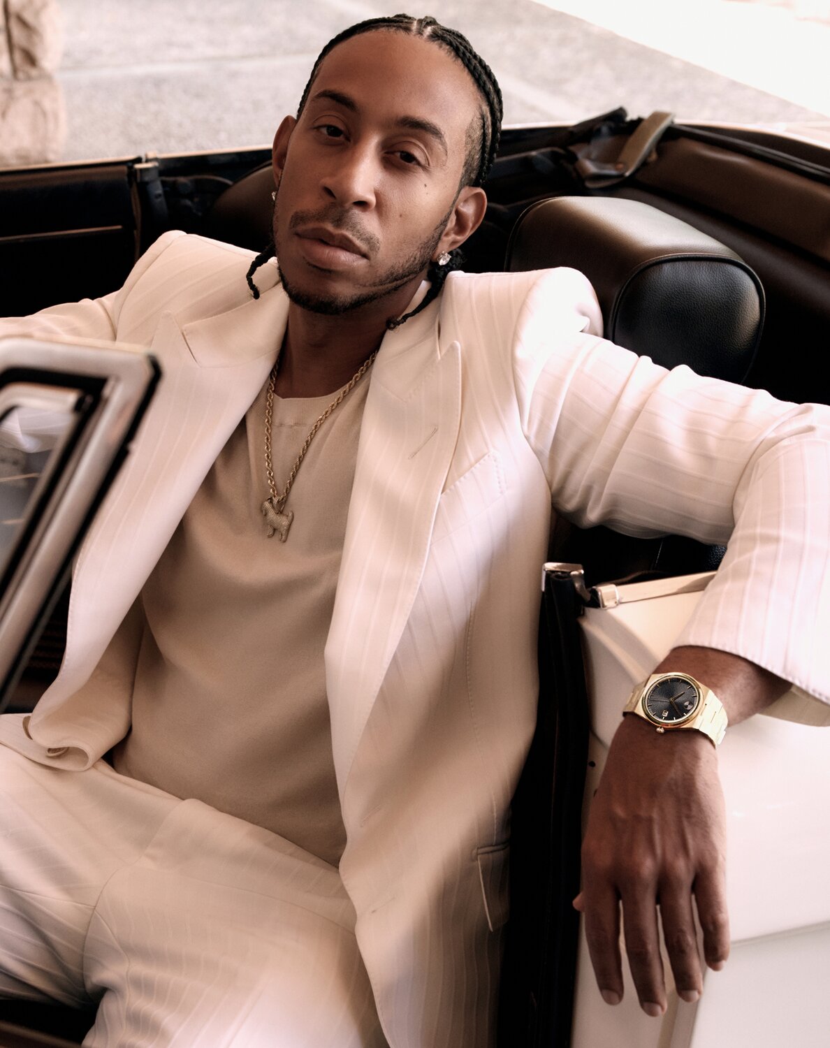 Ludacris sitting in a car, wearing a white suit and the BOLD Quest watch with gold tone bracelet and black dial.