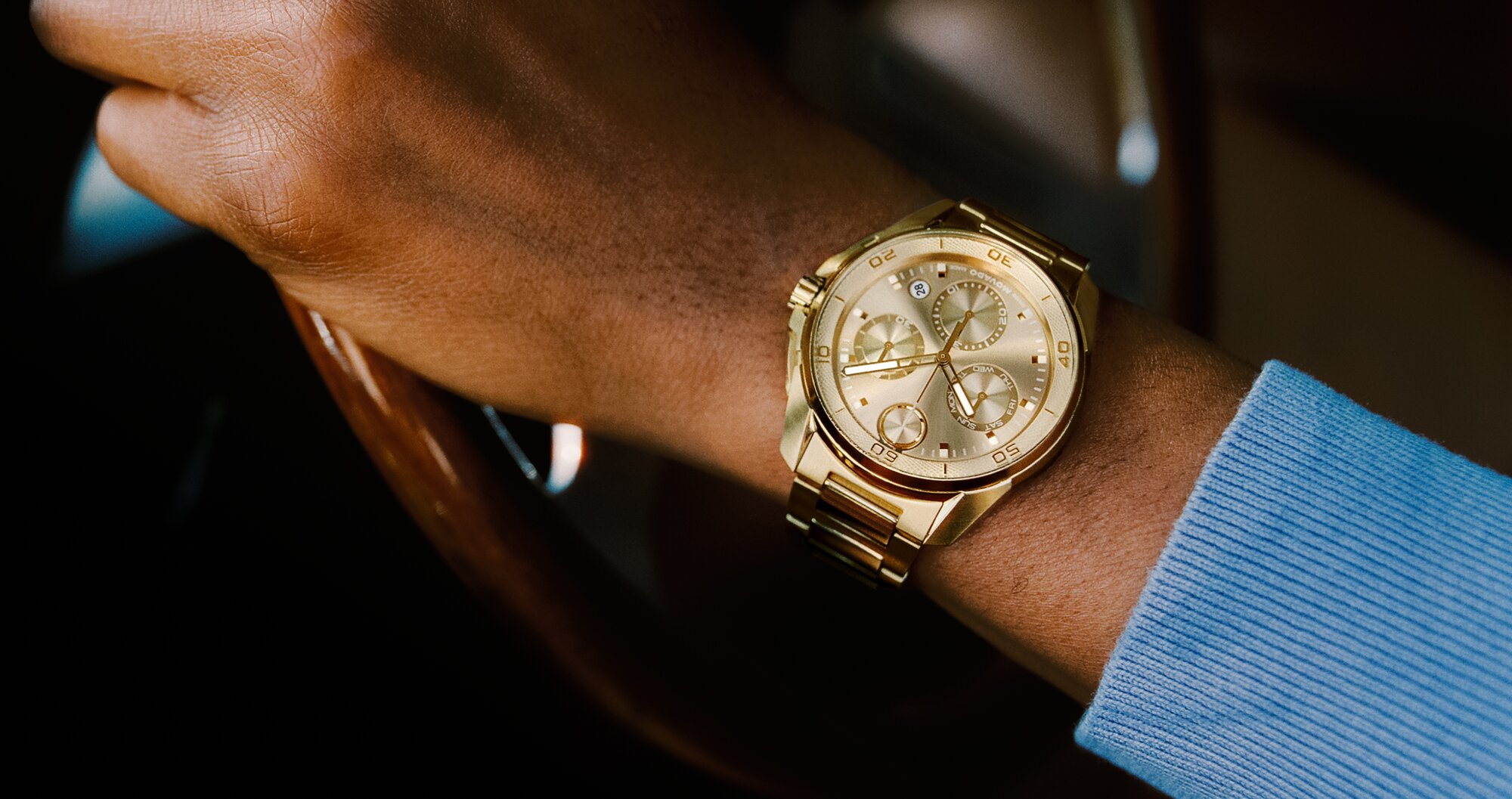 Man in a blue sweater with his hand on a steering wheel of a car wearing the gold tone BOLD Verso S Chronograph bracelet watch.