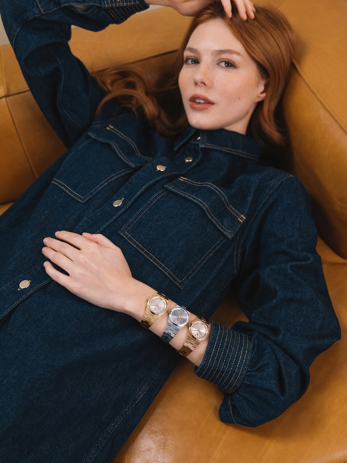 Woman in a denim shirt, laying on a couch, wearing three Movado BOLD Quest Mini bracelet watches in gold tone, rose gold tone and silver.