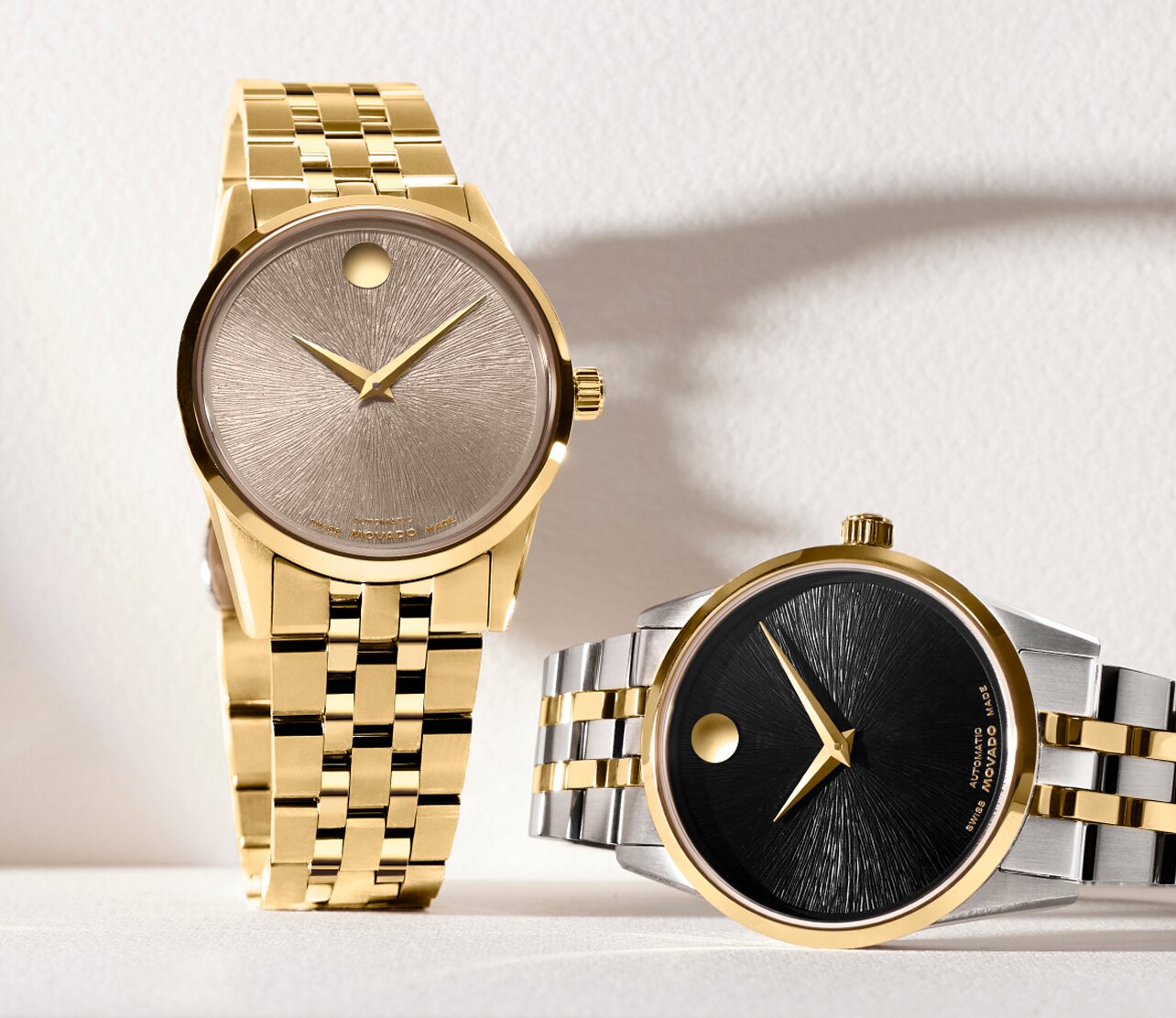 Museum Classic watch with two tone bracelet and black dial laying next to a Museum Classic watch with gold tone bracelet and beige dial.