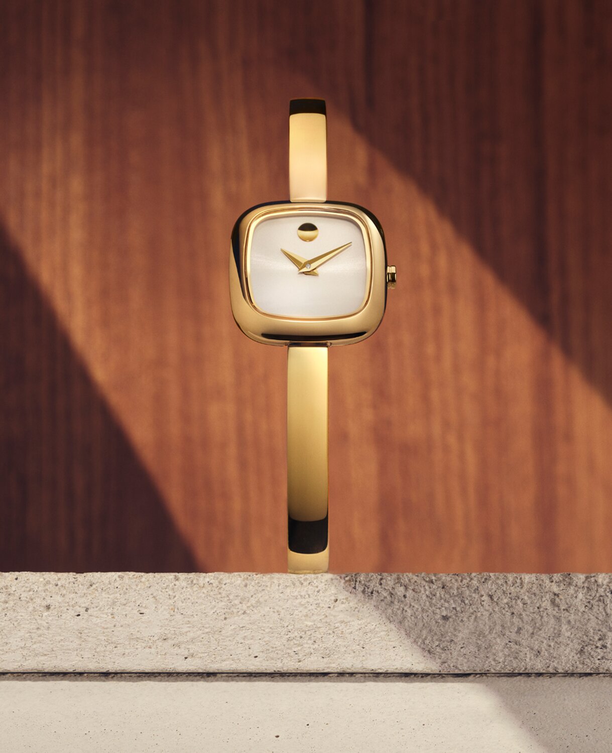Museum Bangle watch with gold tone bracelet and white square dial sitting on stone in front of a wooden panel.
