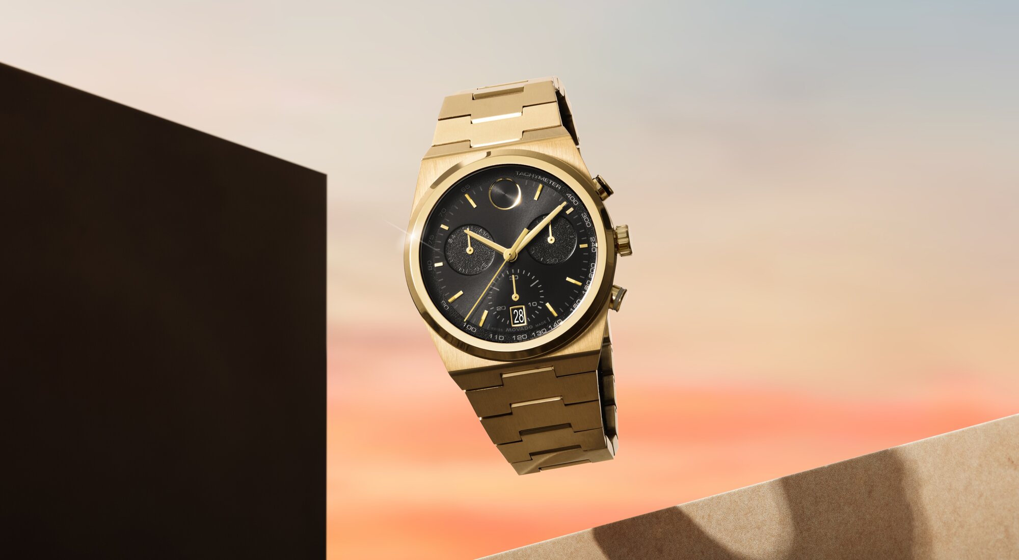 Movado BOLD Quest Chronograph with gold bracelet and black dial floating in front of a sunset.
