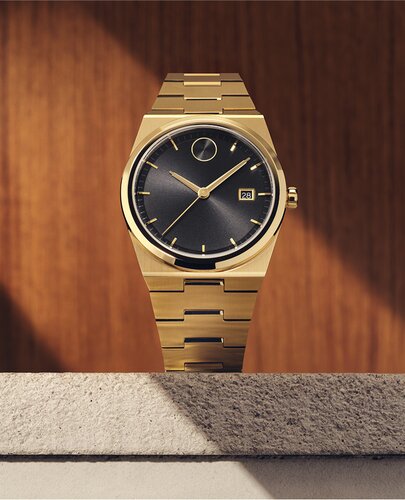 BOLD Quest watch with gold tone bracelet and black dial sitting on stone in front of a wooden panel.