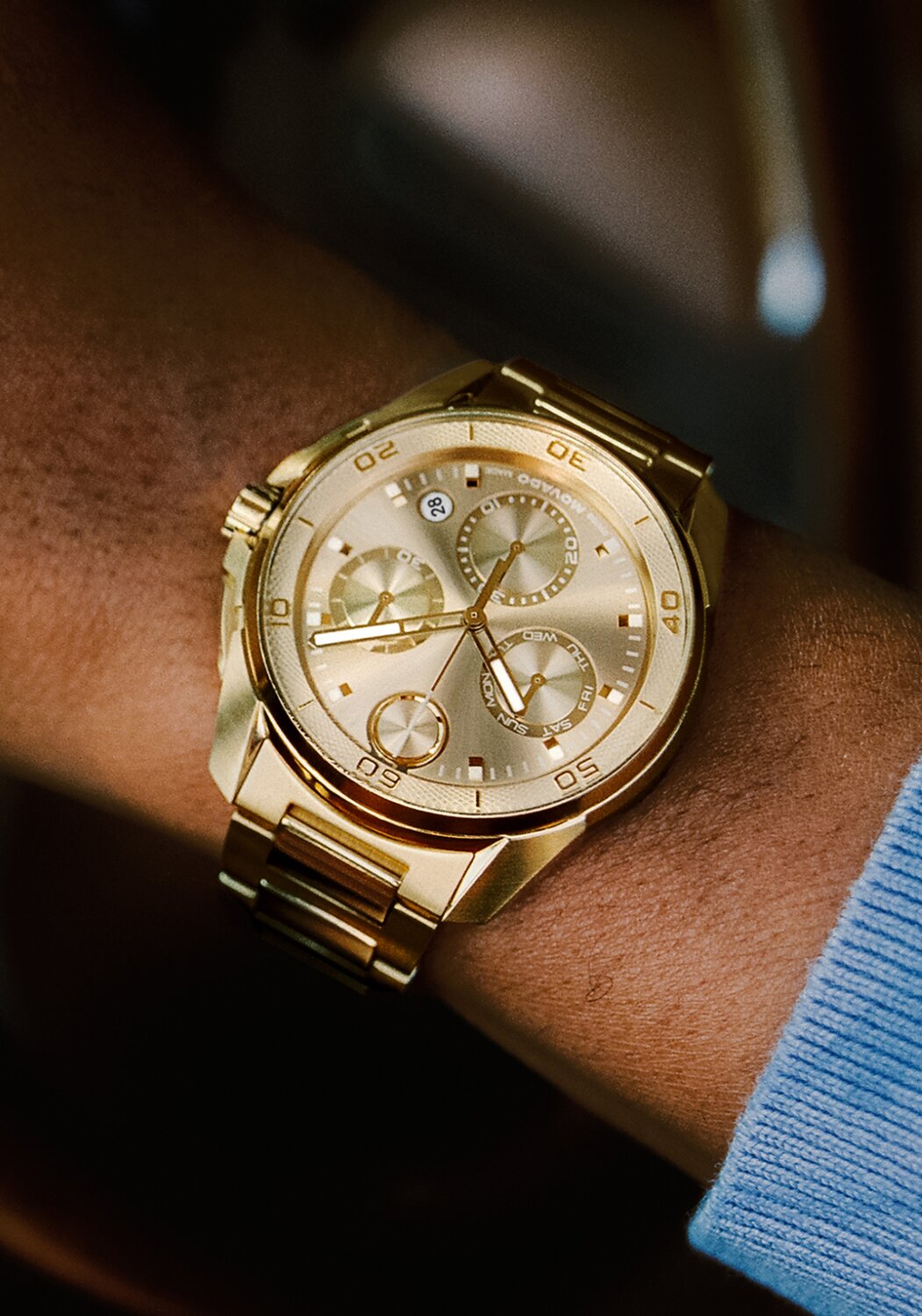 Man in a blue sweater with his hand on a steering wheel of a car wearing the gold tone BOLD Verso S Chronograph bracelet watch.