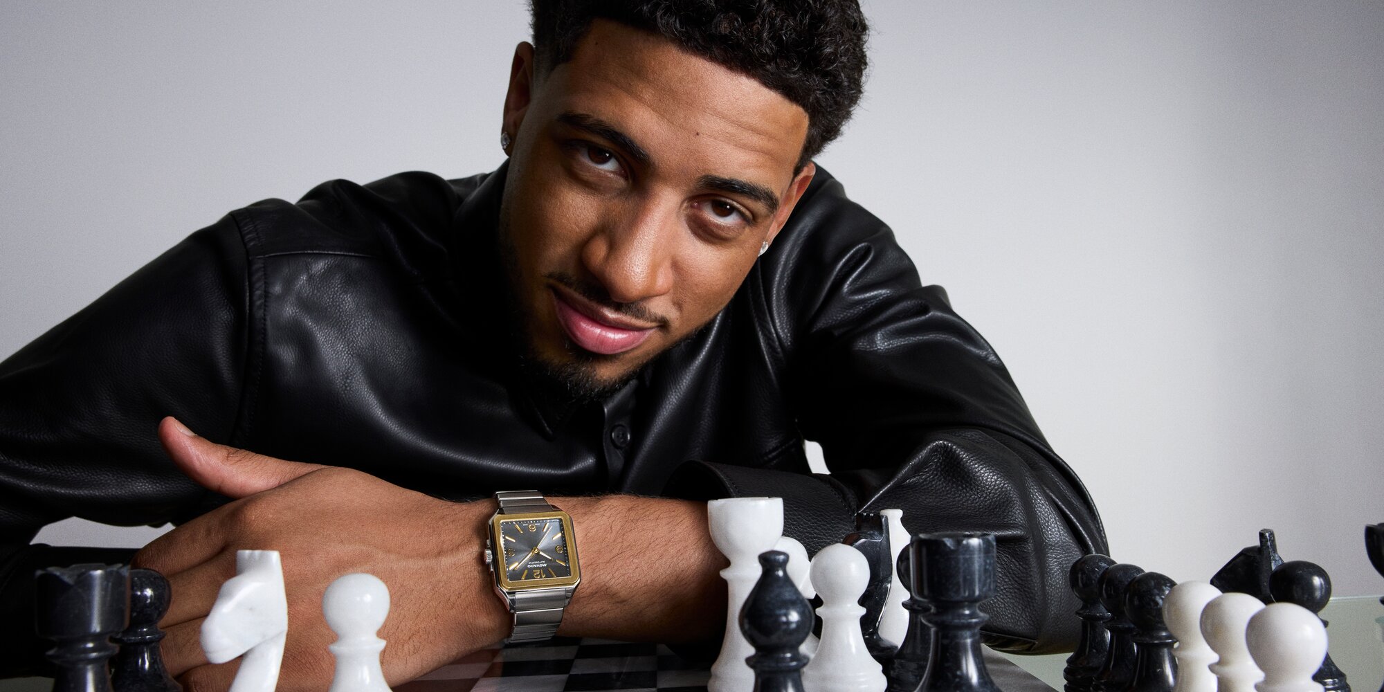 Tyrese Haliburton leaning a chess set in a black leather jacket while wearing the Heritage 1917 watch with stainless steel bracelet and grey dial.