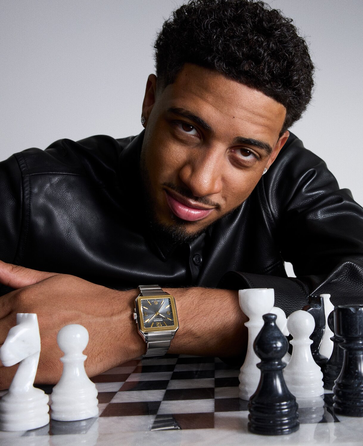 Tyrese Haliburton leaning a chess set in a black leather jacket while wearing the Heritage 1917 watch with stainless steel bracelet and grey dial.