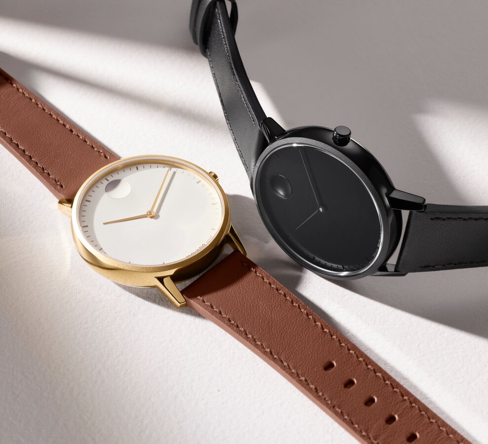 Movado Face watch with brown leather strap and white dial laying next to an all black Movado Face watch with leather strap.