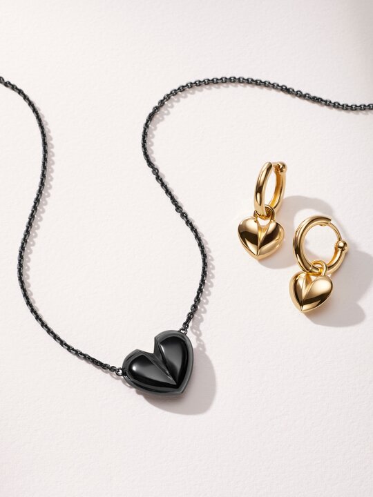 Black plated heart necklace next to gold plated hoop earrings with heart charm.
