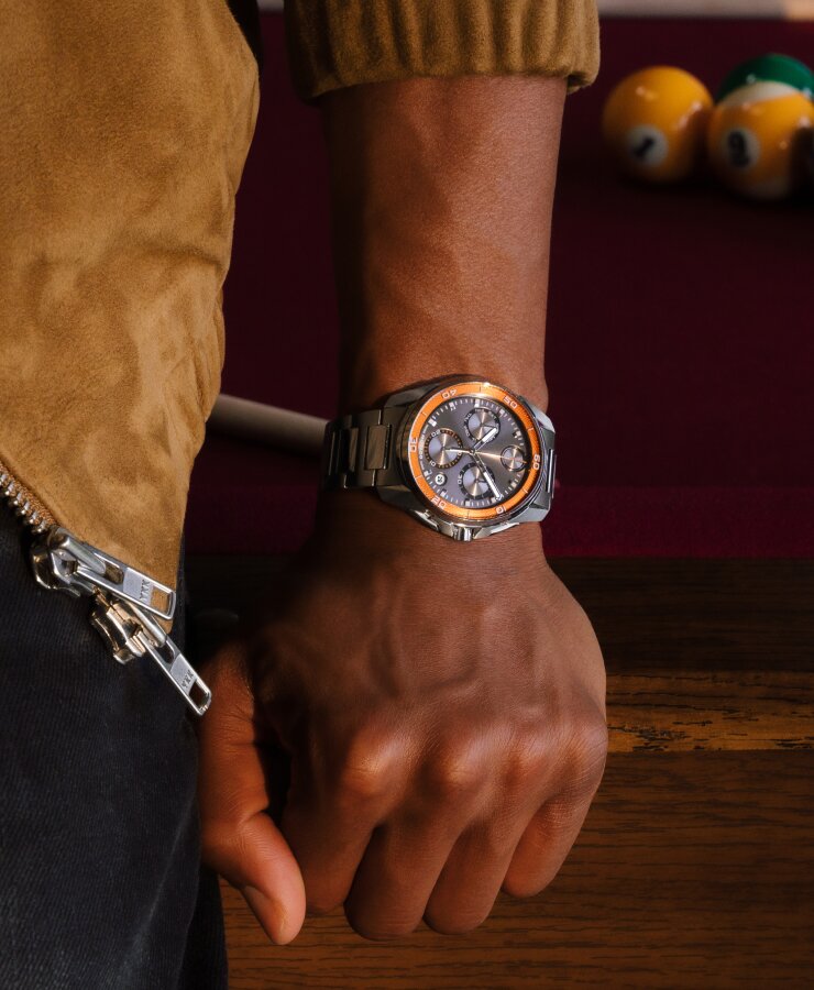 Man kneeling on a pool table wearing the BOLD Verso S Chronograph watch with stainless steel bracelet and grey dial with orange minute track.