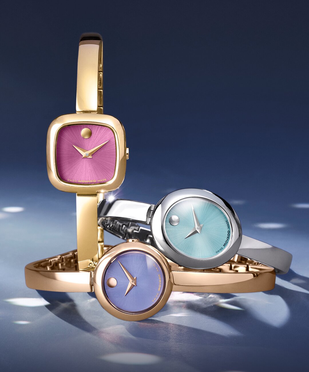 Museum Bangle watch with gold tone bracelet and pink square dial next to two Museum Bangle watches stacked on top of one another. The stainless steel Museum Bangle watch with blue oval dial is laying on top of the rose gold watch with purple dial.
