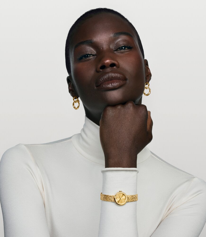 Woman in a white turtle neck wearing gold tone Curve earrings and the gold tone Museum Velura watch with mesh bracelet.