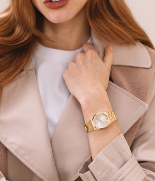 Woman in a pink coat and white t-shirt, holding her collar, wearing the gold tone BOLD Quest Mini bracelet watch.