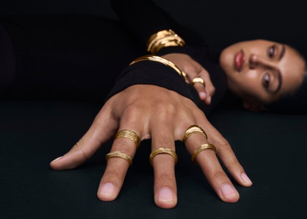Woman laying down with her hand in the foreground wearing 4 gold tone Curve rings.