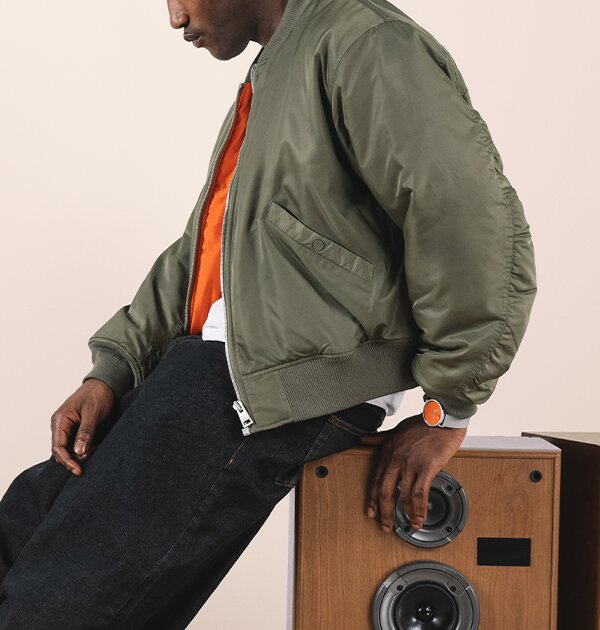Man in a green jacket, leaning on a speaker, wearing a Movado Face watch with grey strap and orange dial 