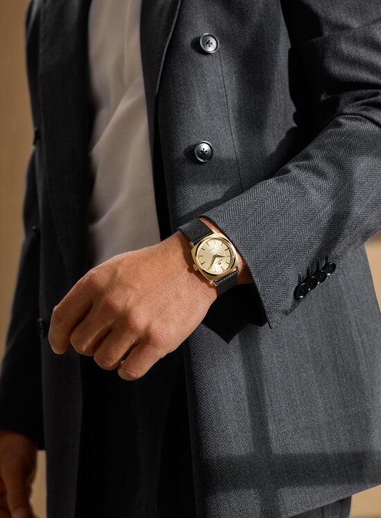 Man in a grey blazer wearing the Heritage Kingmatic watch with black leather strap and gold tone square dial.