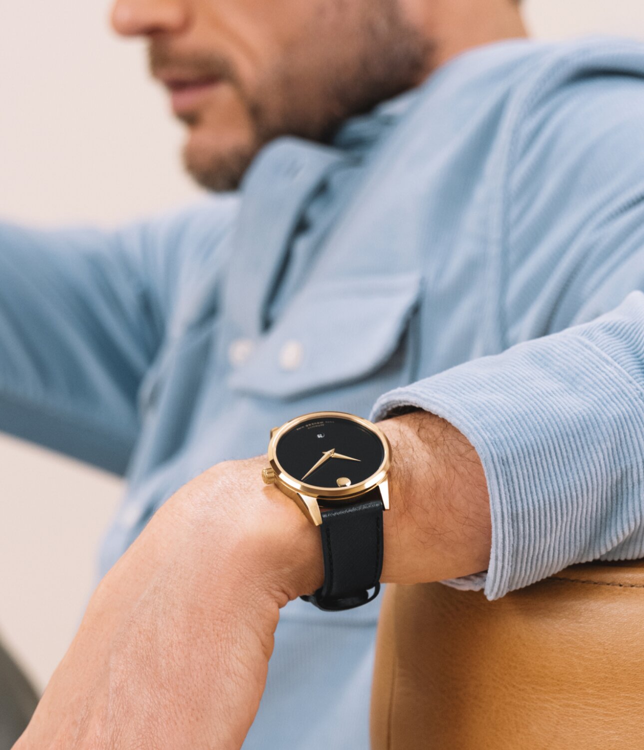 Man in a blue corduroy button down shirt wearing a Museum Classic Automatic watch with black leather strap and black dial with gold tone accents.