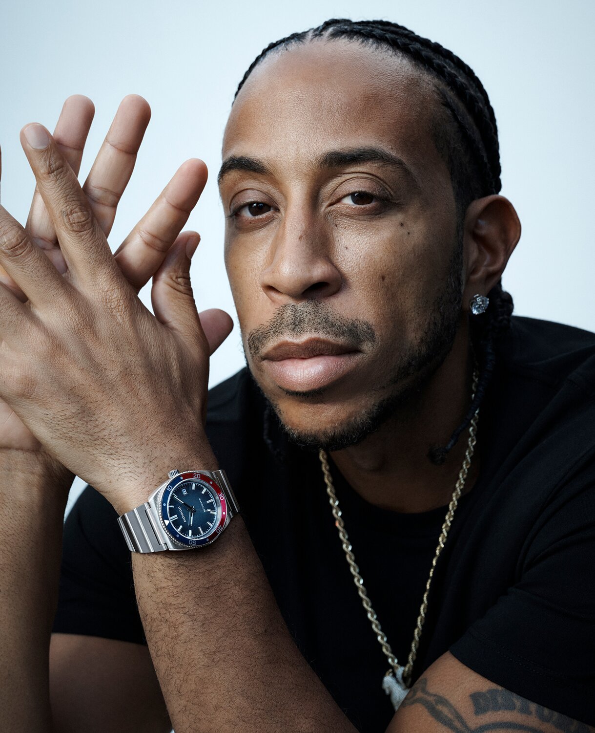 Ludacris in a black t-shirt with his hands crossed wearing an Ondoplan Automatic watch with silver bracelet and blue dial.