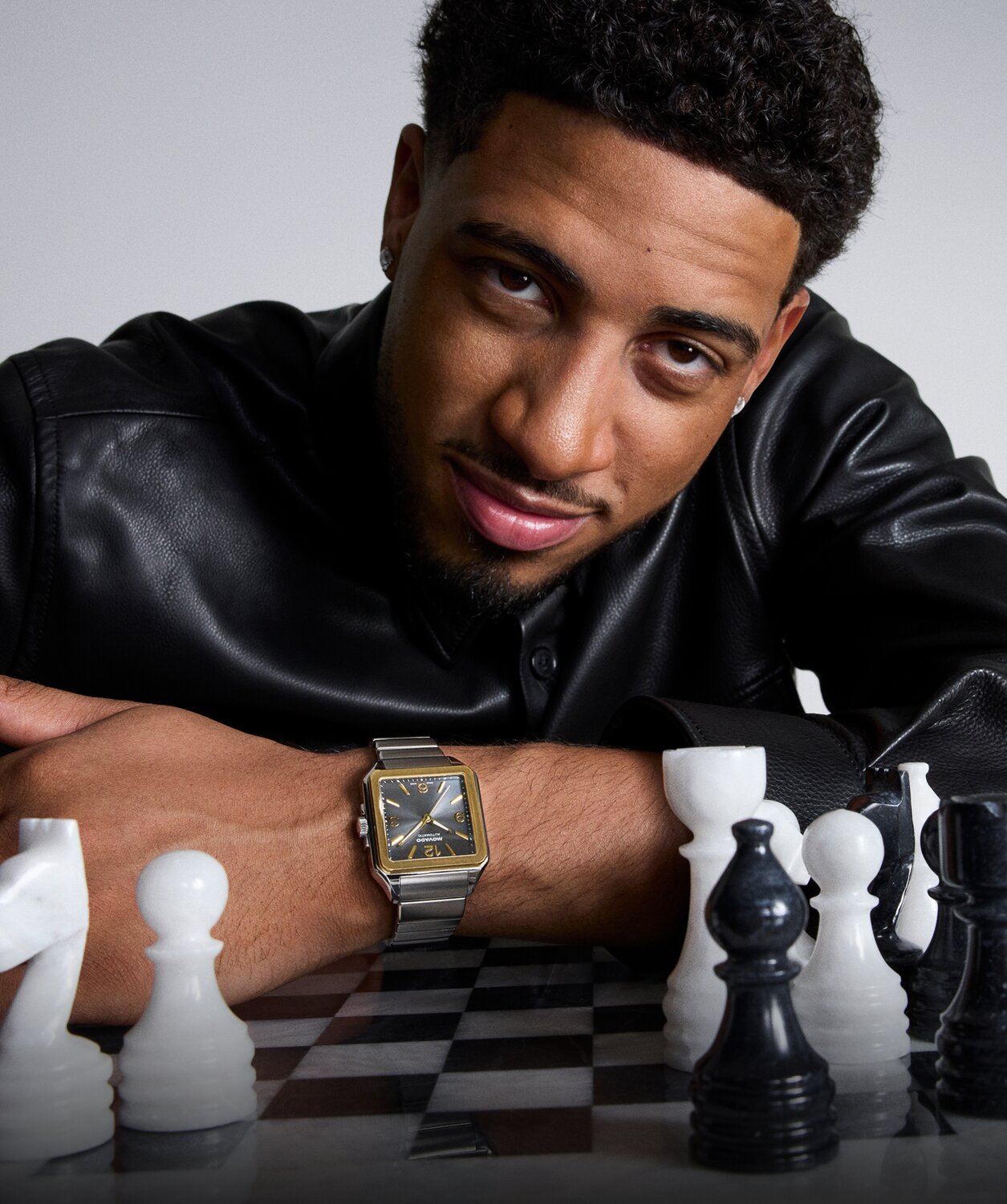 Tyrese Haliburton leaning on a black and white chess board with black in white pieces. He is wearing a black leather button down shirt and the Heritage 1917 stainless steel bracelet watch and square grey dial.