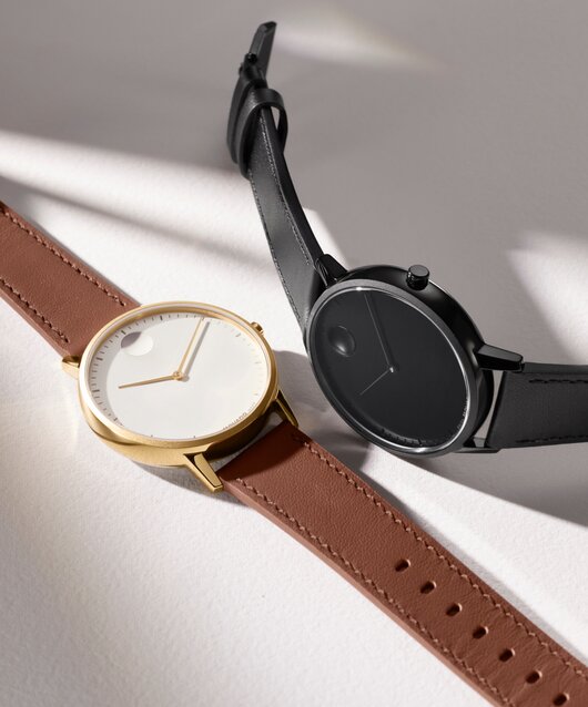 Movado Face watch with brown leather strap and white dial laying next to an all black Movado Face watch with leather strap.
