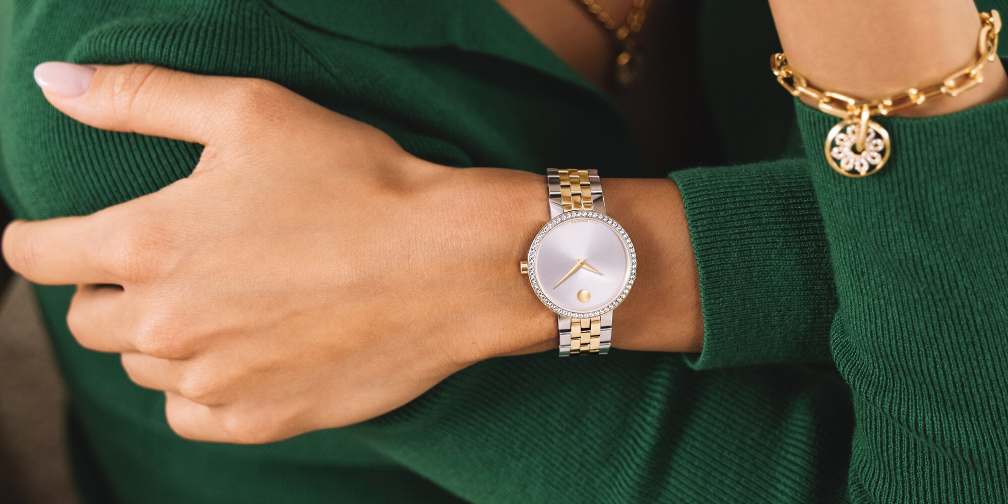 Woman in a green sweater wearing a two tone Museum Classic bracelet watch featuring lab grown diamonds and a gold tone fleur necklace and bracelet that features lab grown diamonds.