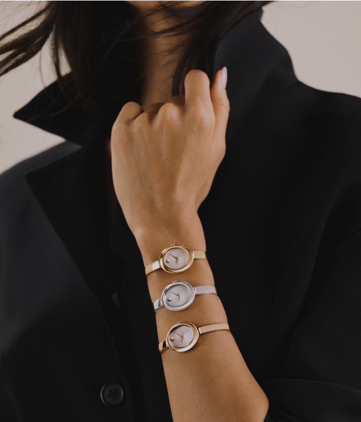 Woman in a black coat wearing a stack of three Oval Museum Bangles on her arm. Leading with gold tone, followed by silver, and ending with rose gold tone.