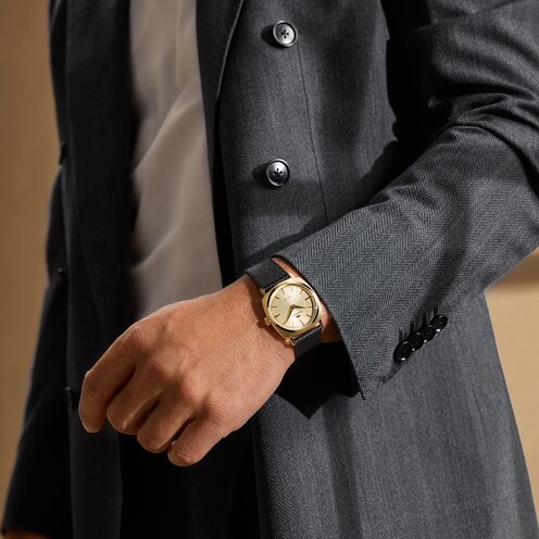 Man in a grey blazer wearing the Heritage Kingmatic watch with black leather strap and gold tone square dial.
