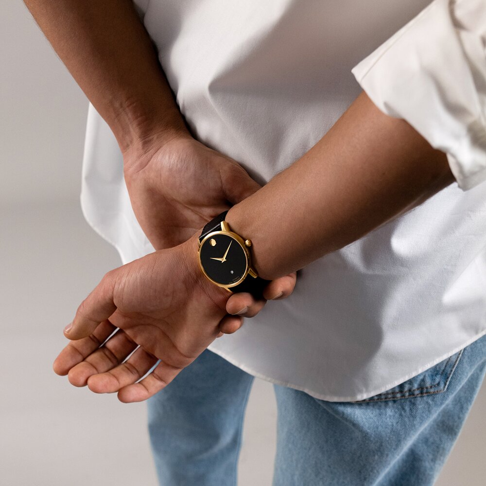 Man in a white top and jeans with their hands crossed behind their back wearing the Museum Classic watch with black leather strap and black dial.