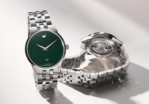 Front of a Museum Classic silver bracelet  watch with green dial behind the back of the watch with a transparent case back that showcases the automatic movement