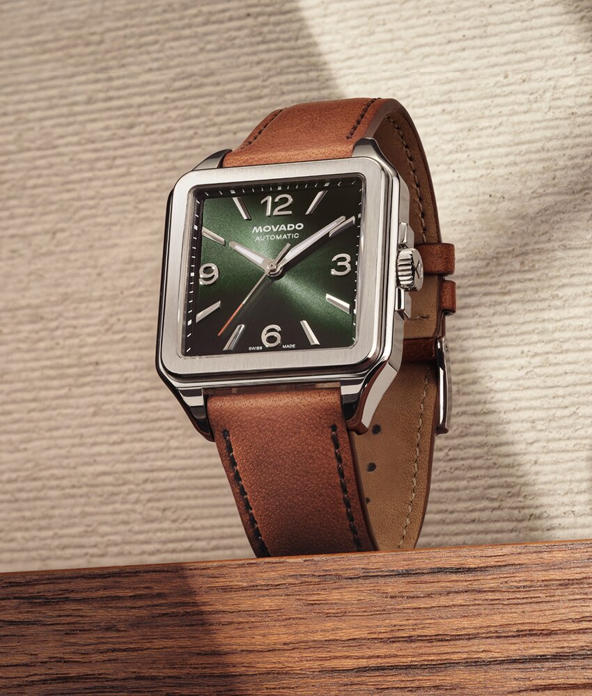 Heritage 1917 watch with brown strap and green dial sitting on wood in front of an ivory textured background.