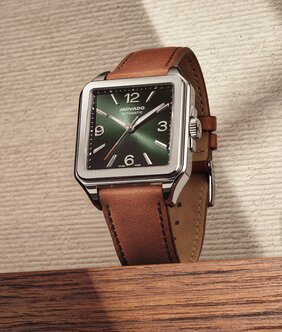 Christian McCaffrey's hand in the pocket of his white pants while wearing a grey Heritage 1917 watch with brown strap and green dial sitting on wood in front of an ivory textured background.