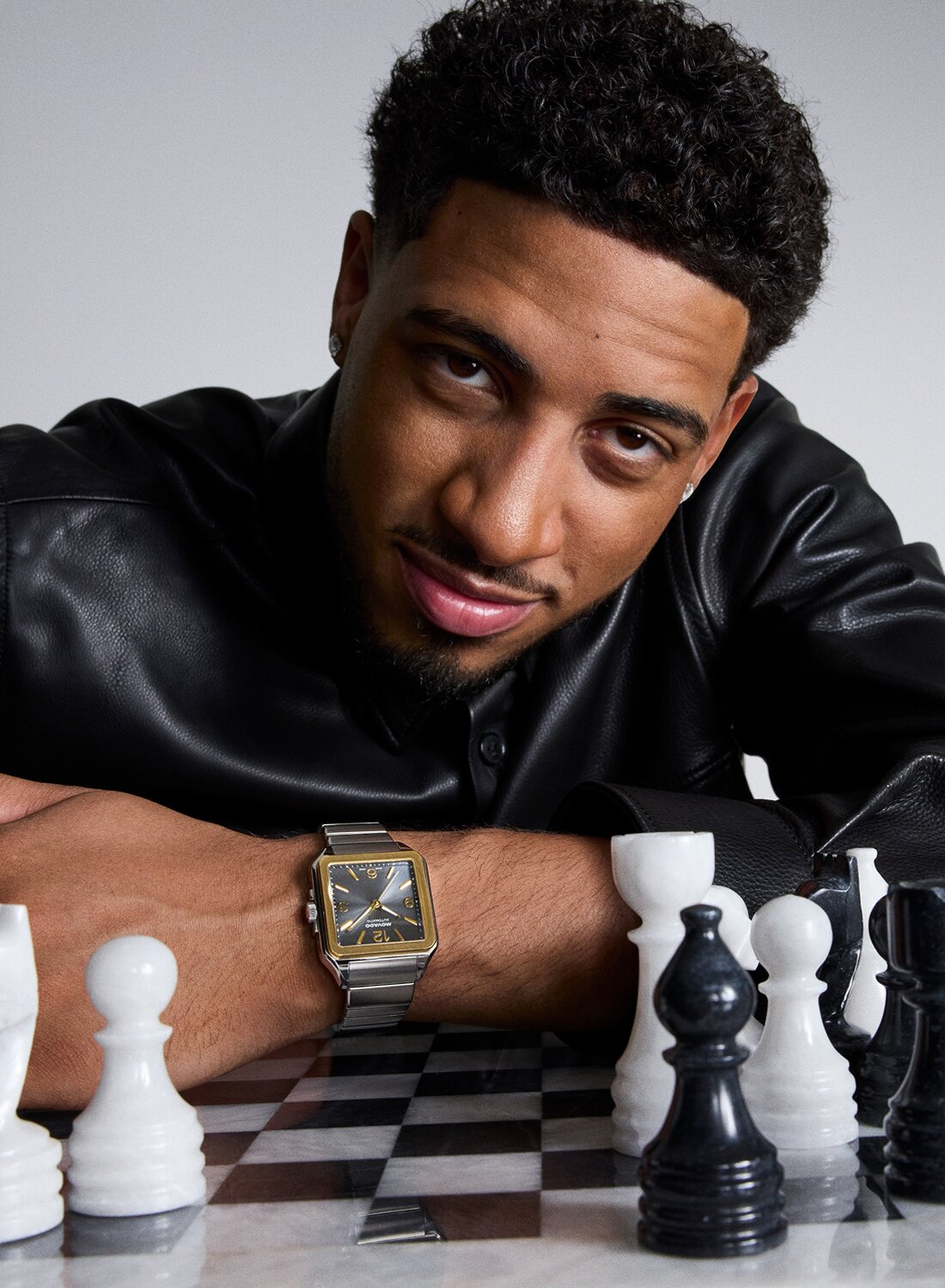 Tyrese Haliburton leaning a chess set in a black leather jacket while wearing the Heritage 1917 watch with stainless steel bracelet and grey dial.