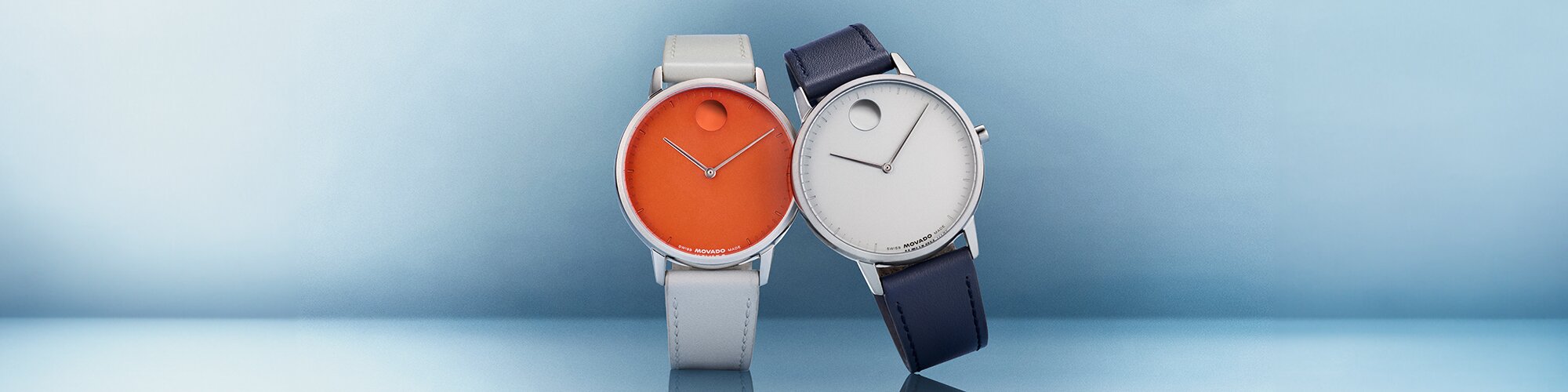 Silver strap Movado Face watch with orange dial next to a blue leather strap Movado Face watch