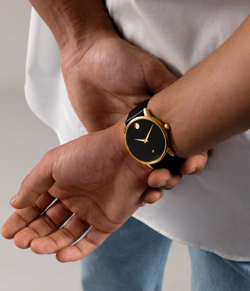 Man with hands behind his back wearing the Museum Classic Automatic watch with black strap and black dial with gold accents.