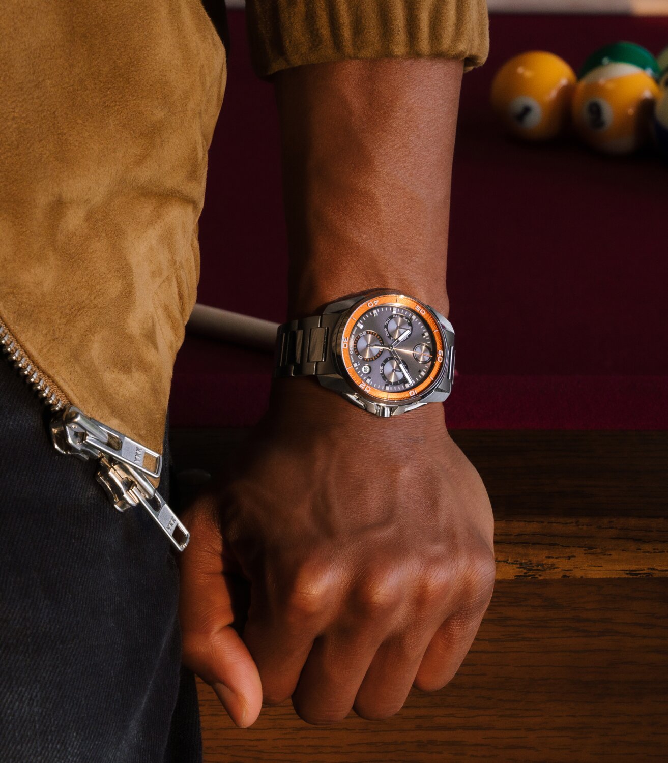 Man kneeling on a pool table wearing the BOLD Verso S Chronograph watch with stainless steel bracelet and grey dial with orange minute track.