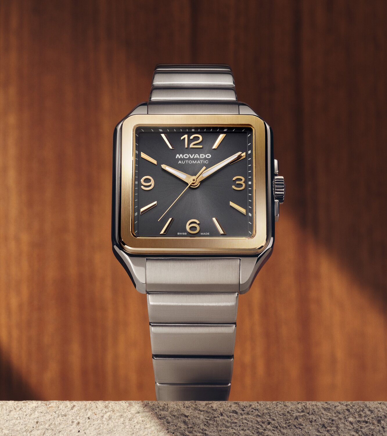 Heritage 1917 with stainless steel bracelet and square grey dial sitting on concrete in front of a wooden wall.