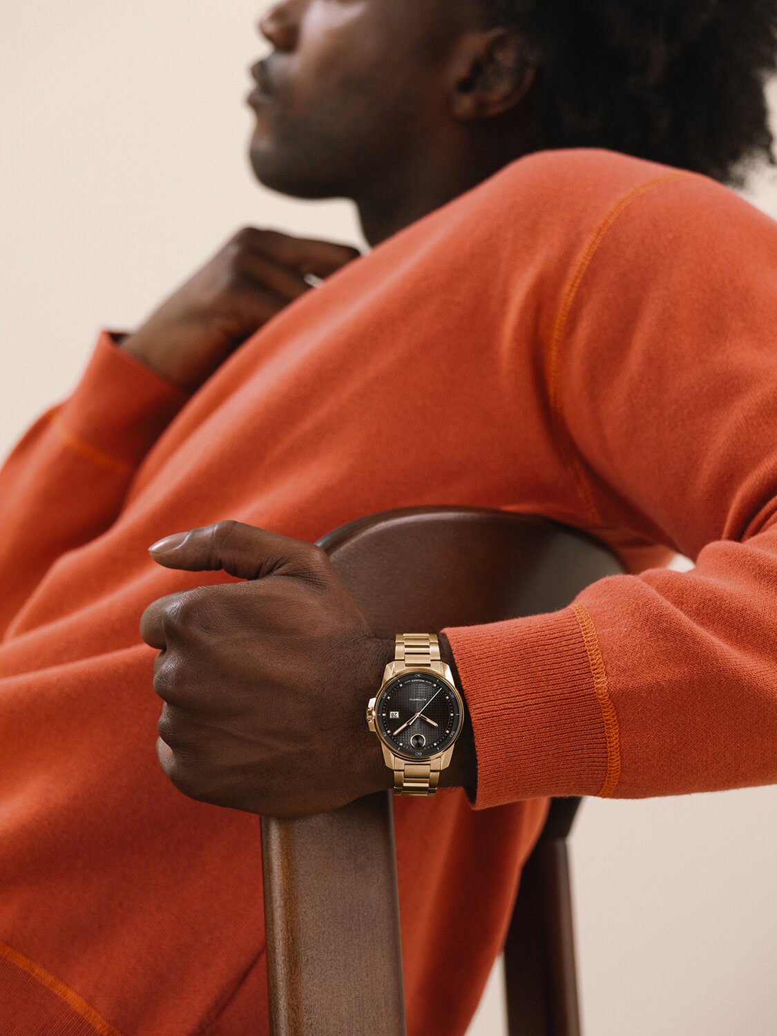 Man in an orange sweater, sitting in a chair, wearing a BOLD Verso Automatic watch with goldtone bracelet and grey dial.